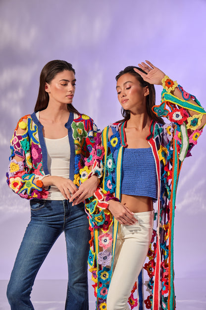 Two women modeling vibrant, multicolored crochet cardigans with floral and geometric patterns, set against a soft purple studio backdrop.