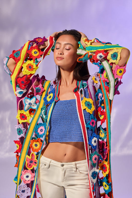 A woman wearing a vibrant, multicolored open-front crochet cardigan with large flower motifs, styled over a blue knit crop top and cream pants, set against a soft purple background.